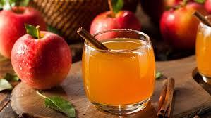 A rustic wooden table displaying a pitcher of unpasteurized apple cider, glasses filled with cider, and a basket of fresh apples, with autumn leaves scattered around.